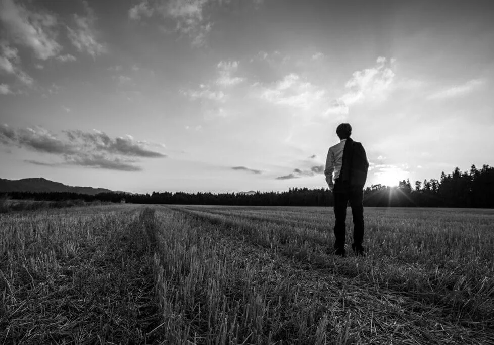 Businessman in elegant suit with his jacket hanging over his shoulder standing in mown wheat field looking into the distance under a majestic evening sky with a setting sun.
