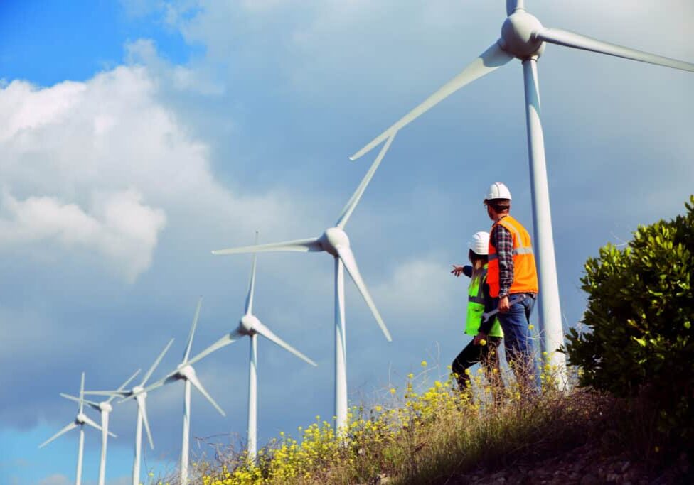 Two young caucasian technician engineer man and woman wearing white worker headwear standing, working, checking farm field system and looking up at wind turbine farm power generation station in rural landscape.